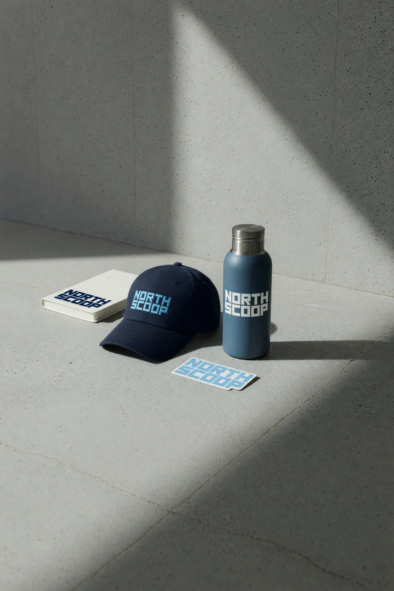 A navy cap, a steel bottle, a notebook and a sticker printed with a North Scoop logo on a concrete floor in hard sunlight.