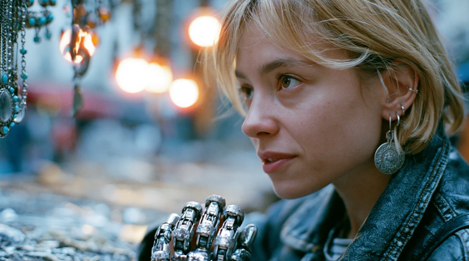 A young woman with short blond hair and a metal robotic hand browses jewelry at a market stall.