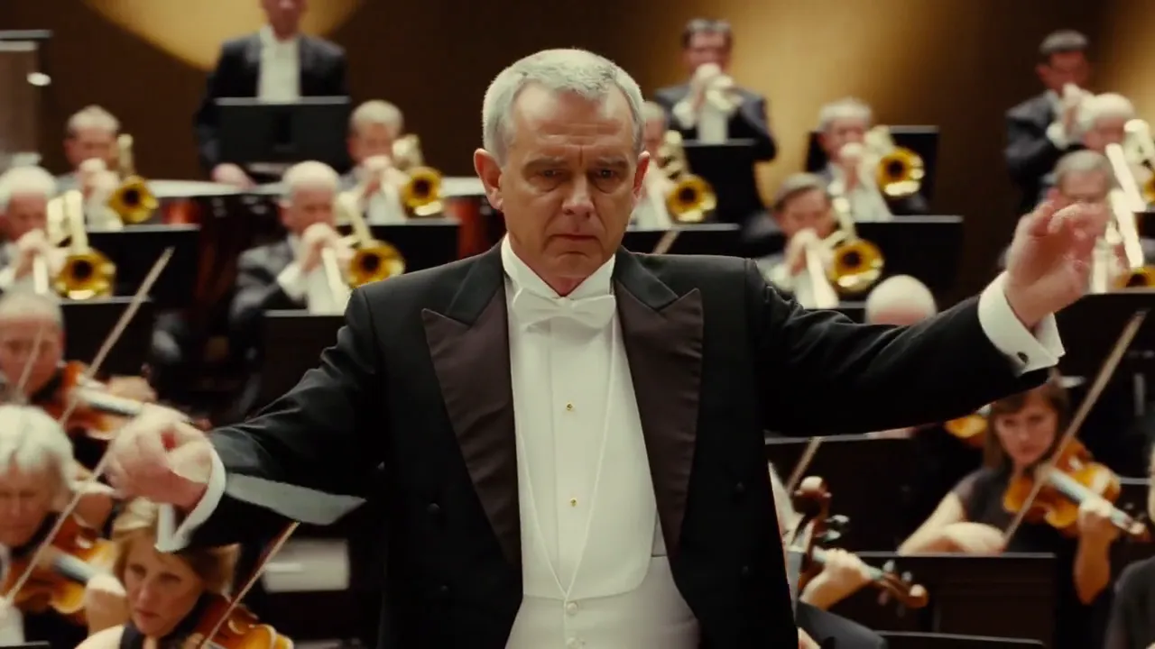 A conductor raises his baton in front of a full orchestra in a concert hall.