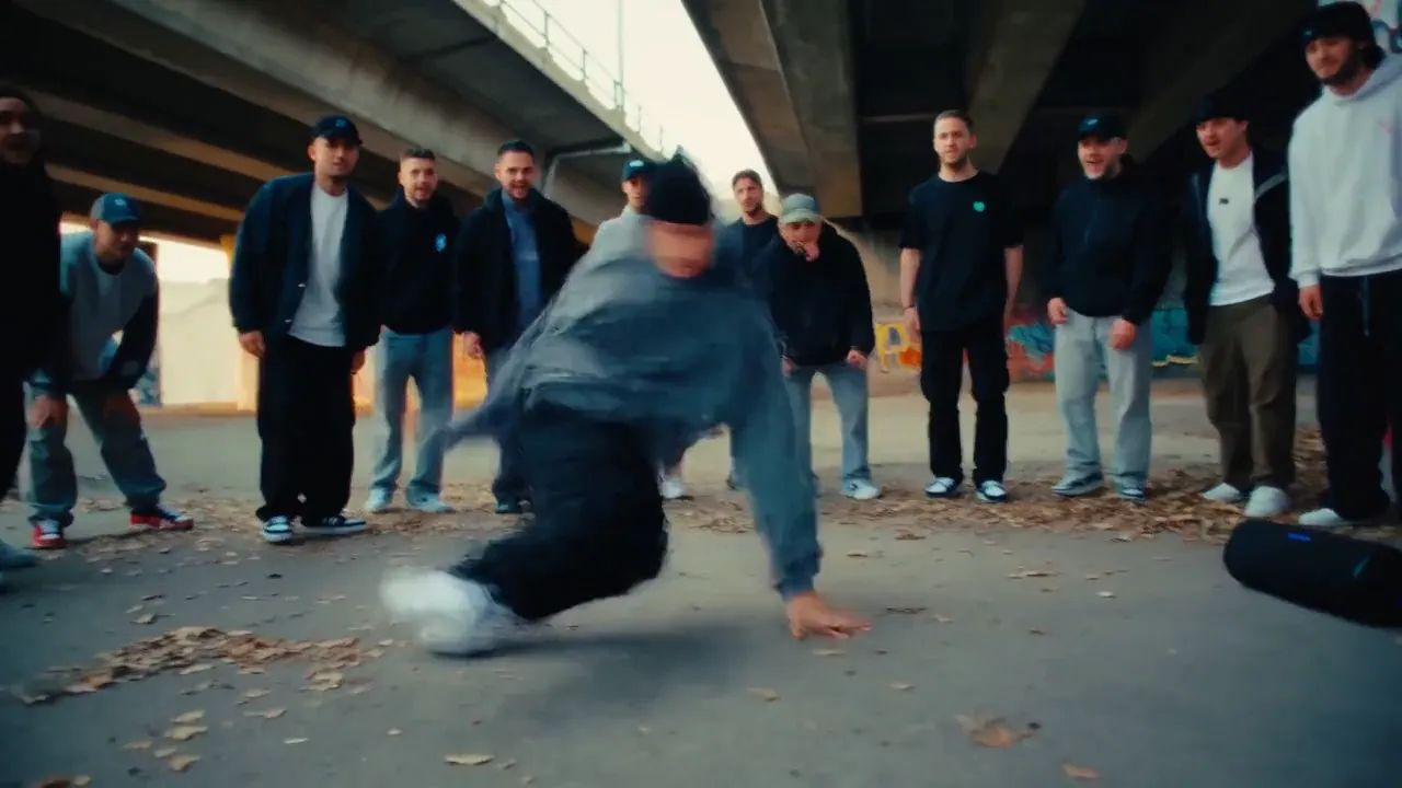 A breakdancer spins on the concrete under a highway overpass while a crowd watches.