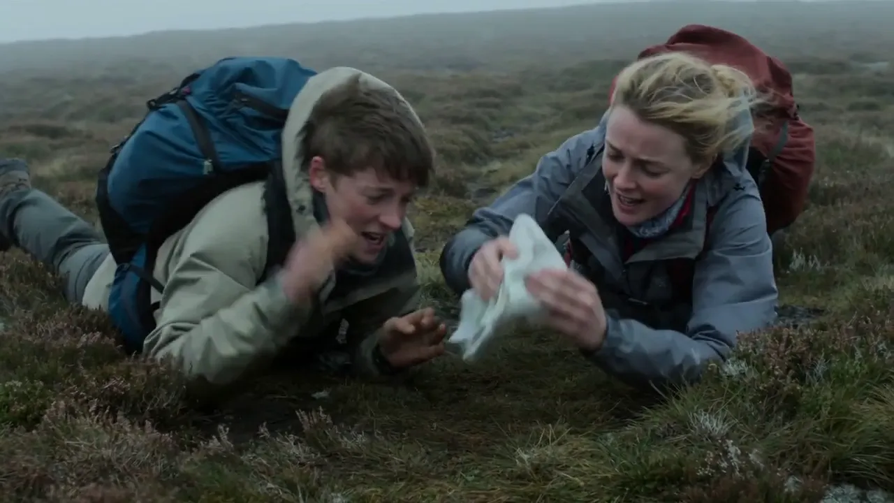 Two hikers lie laughing in the heather as they catch a map on a misty moor.