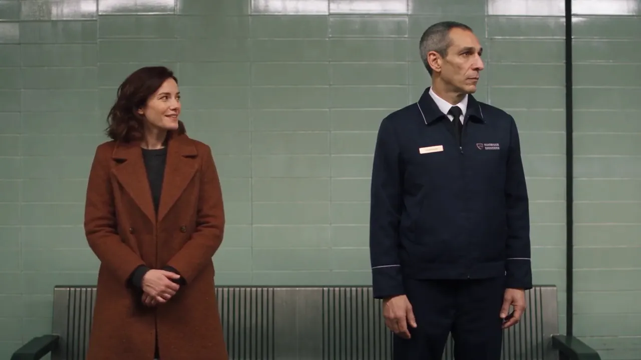 A woman in a brown coat smiles at a uniformed station worker on a subway platform.