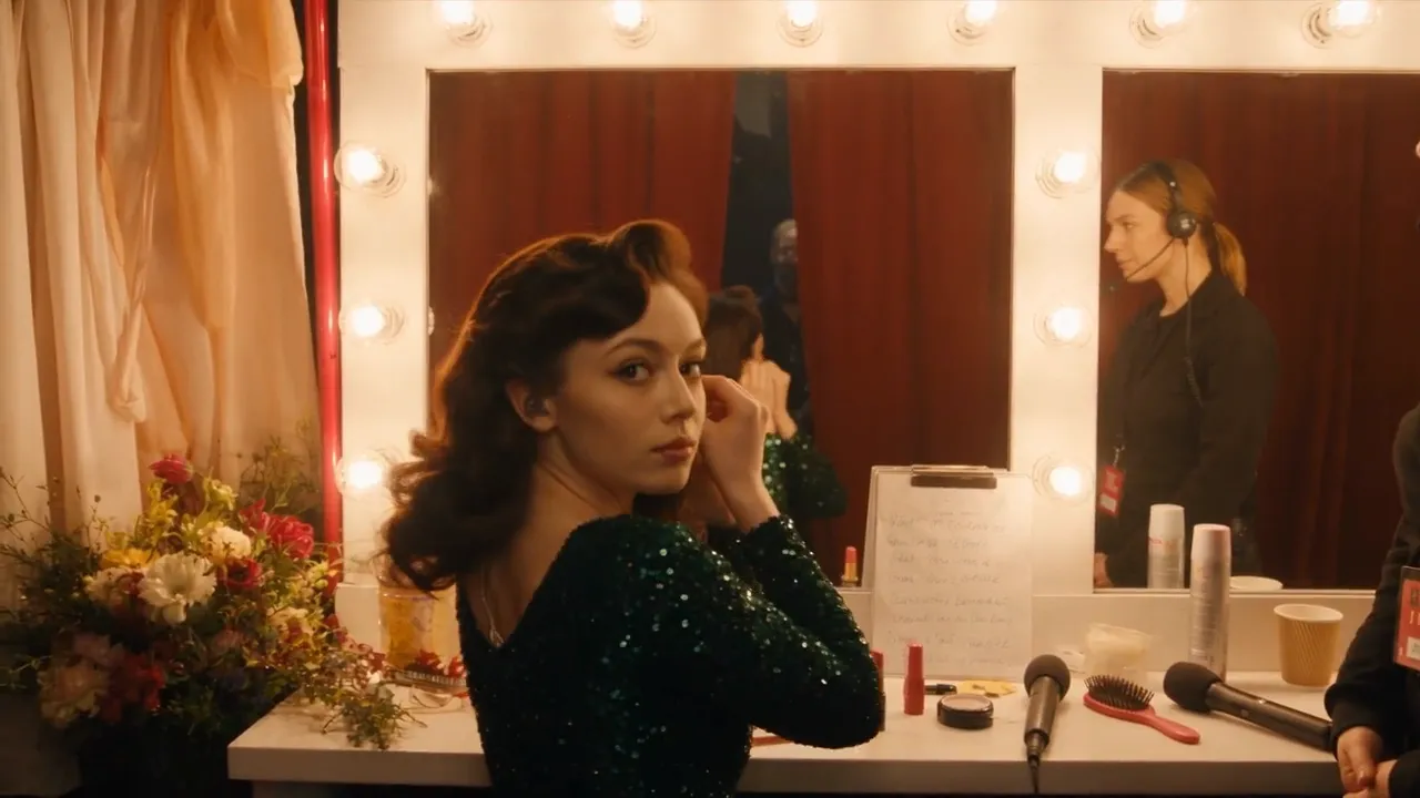 A singer in a green sequined dress glances back from a backstage makeup mirror.
