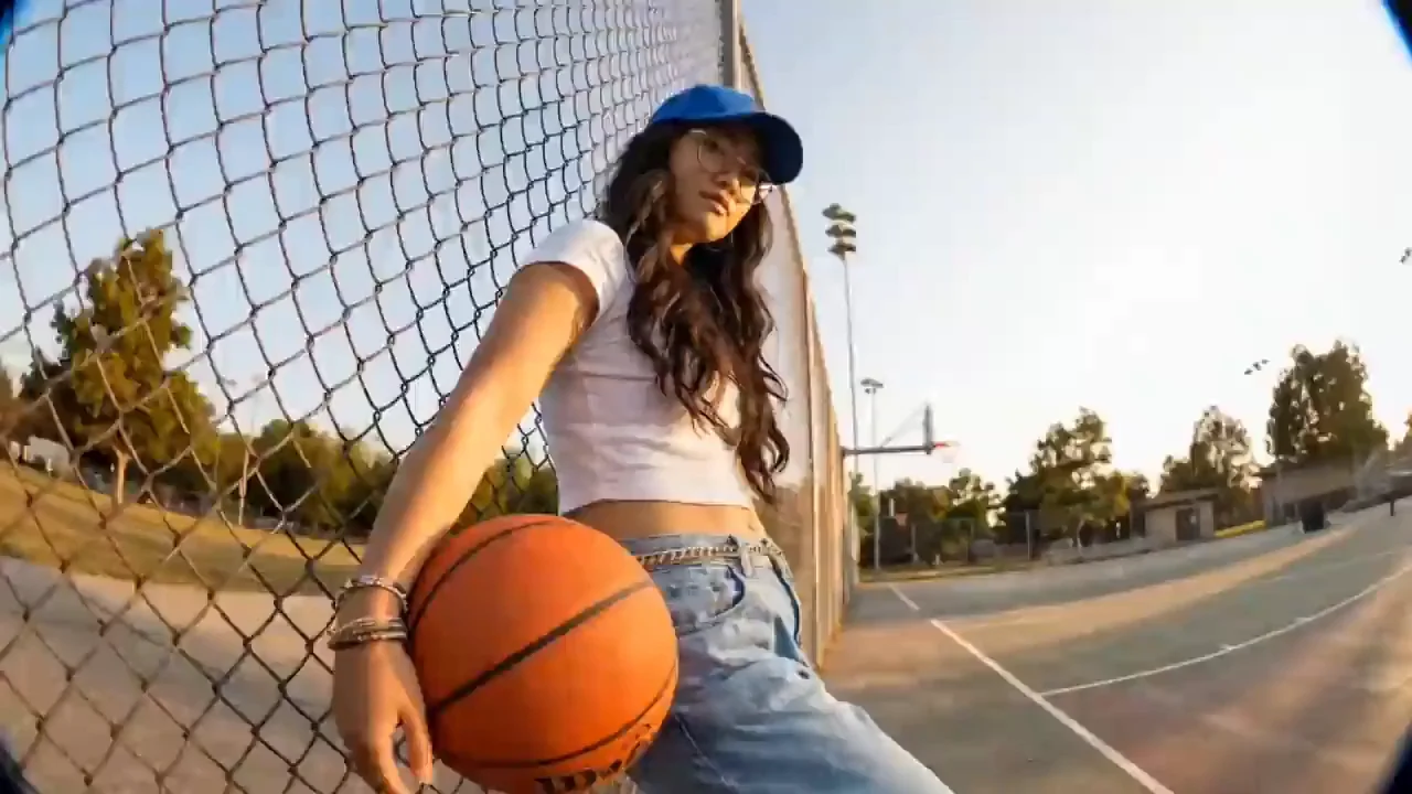 A young woman in a blue cap dribbles a basketball on an outdoor court at sunset.